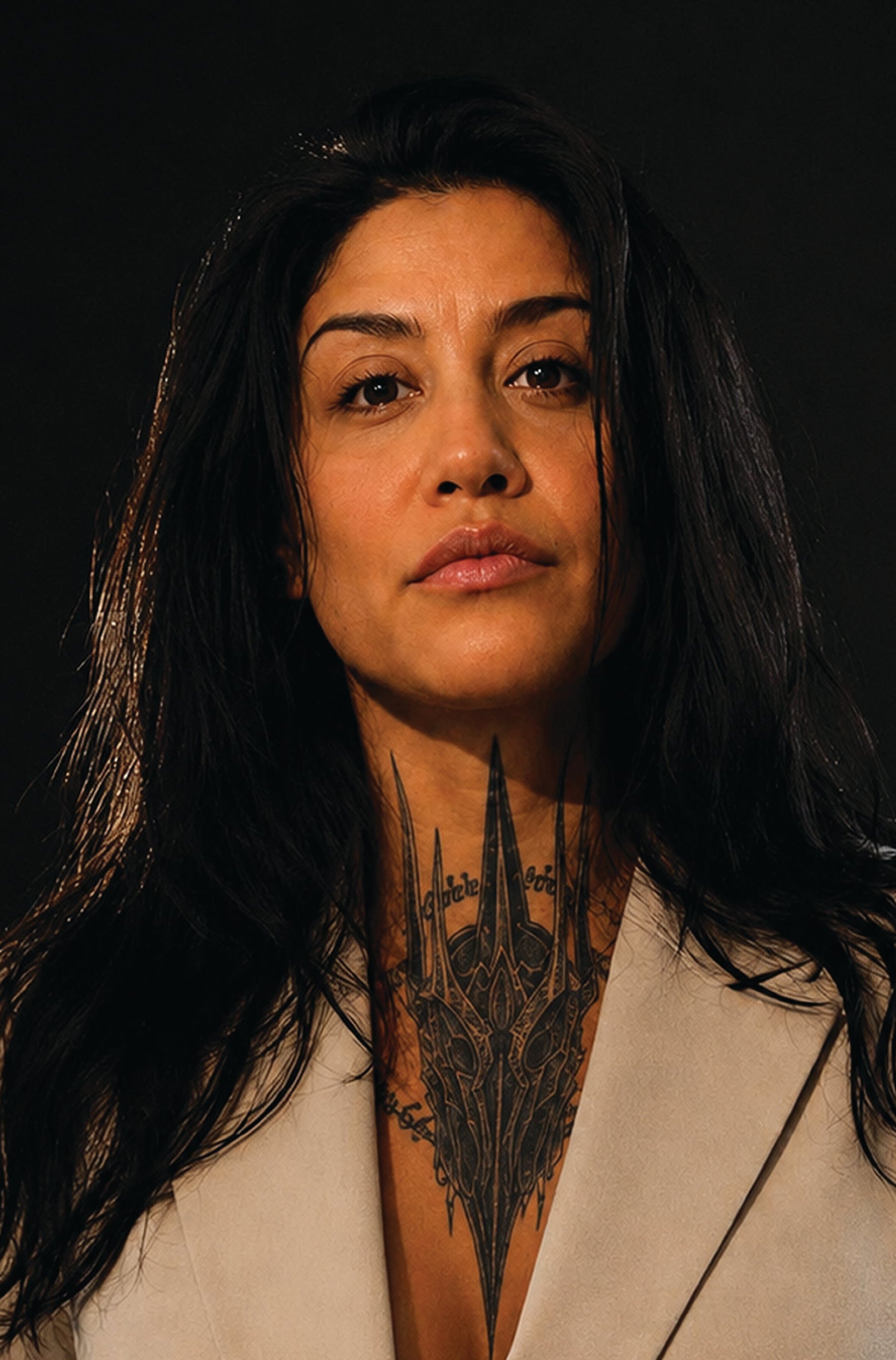 A close-up portrait of a woman with long dark hair and a detailed black gothic-style tattoo on her neck, wearing a light beige blazer against a dark background.